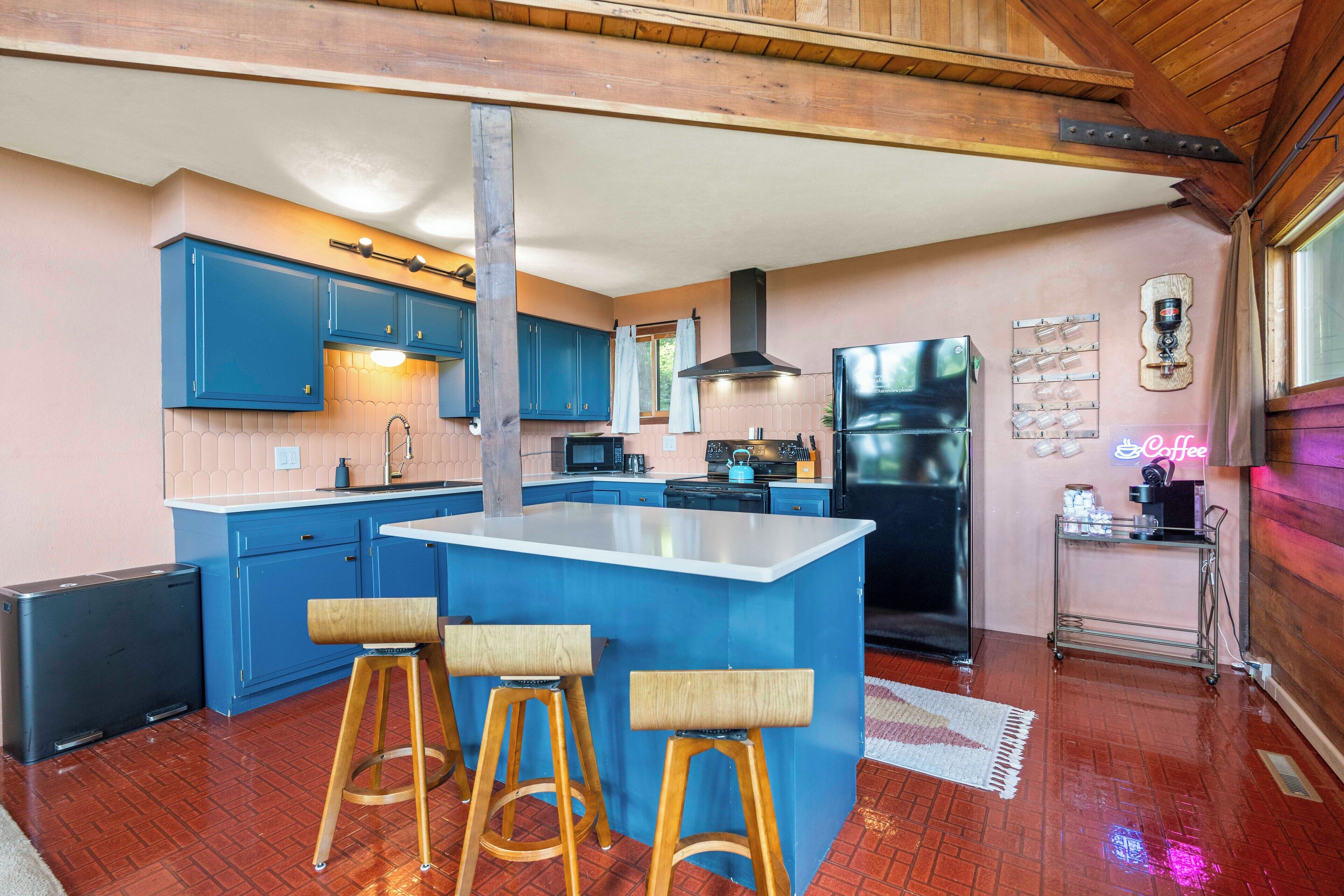 A kitchen with blue cabinets, a central island with stools, a black refrigerator, and a neon sign.