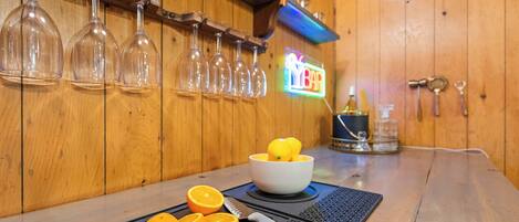 A bar area with wine glasses on a rack, a bowl with lemons, and a cutting board with orange slices.