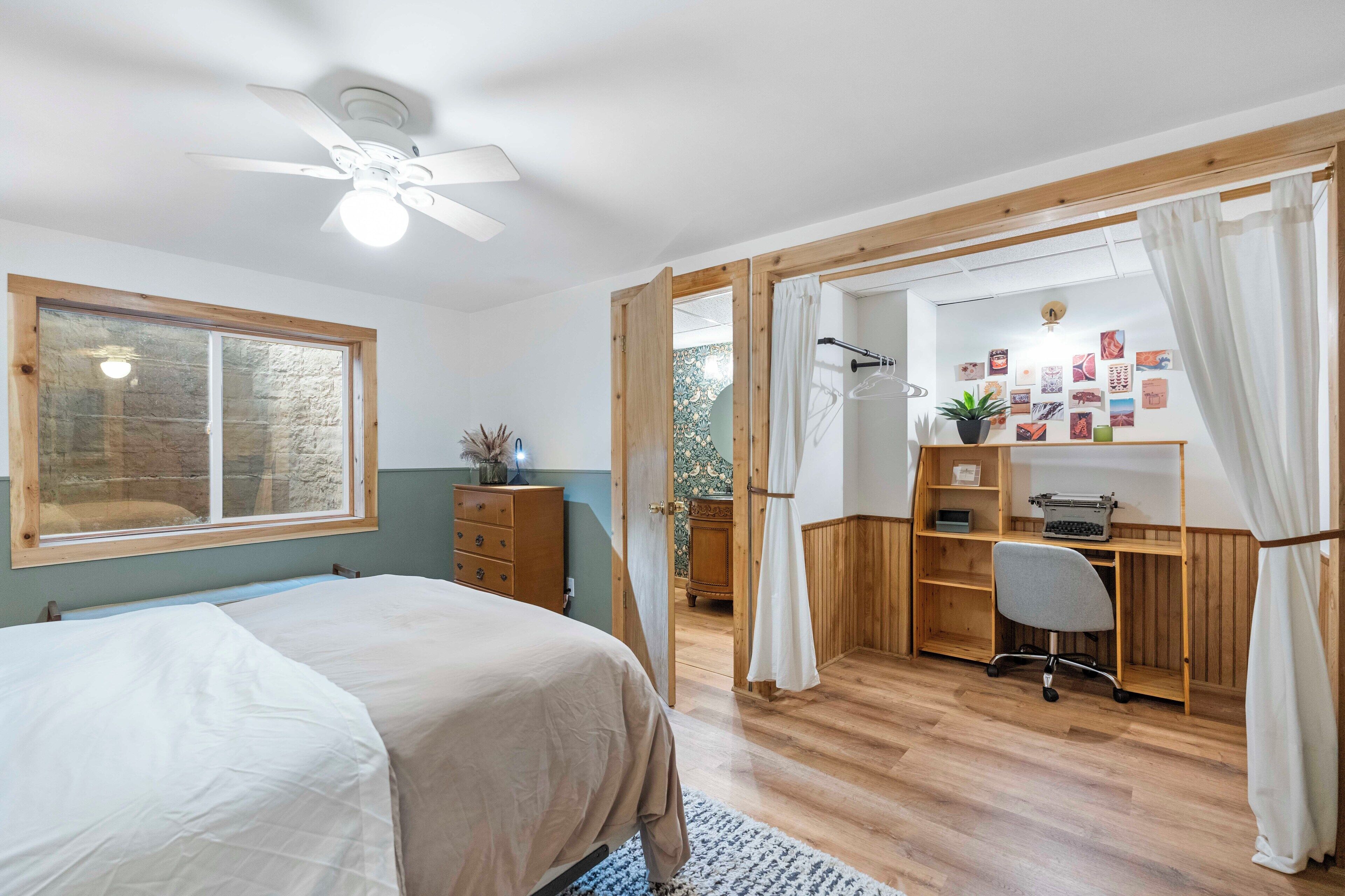 Une chambre à coucher comprenant un lit, un bureau avec une chaise, un ventilateur de plafond et une fenêtre avec des rideaux.