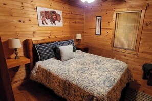 Ein Schlafzimmer einer Holzhütte mit einem Bett, zwei Nachttischen, einer Lampe, einem Bilderrahmen mit einem Bison und einem Fenster mit Jalousien.