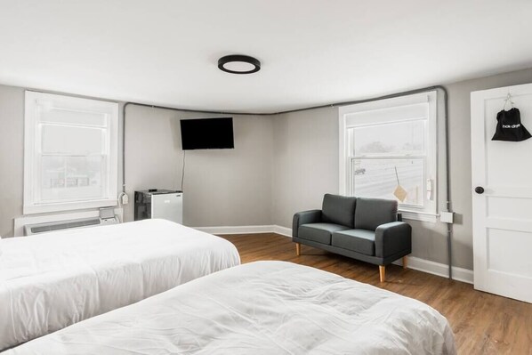 A modern hotel room with two beds, a gray sofa, a flat-screen TV, and a view of the city through large windows.