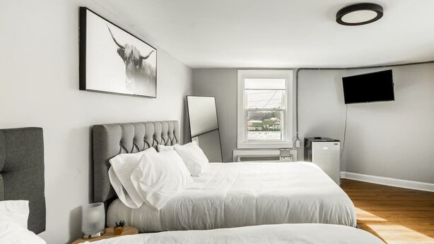 Ein modernes Schlafzimmer mit zwei Betten, einem grauen Kopfteil, einem an der Wand montierten Fernseher und einem Fenster mit Blick auf Gebäude.