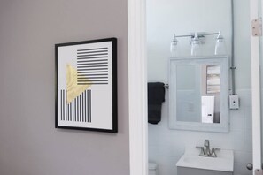Une salle de bain avec une œuvre d’art abstraite encadrée, un miroir et un lavabo blanc.
