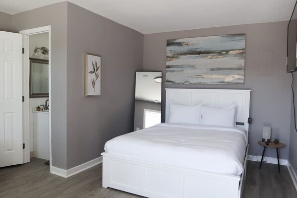 Un dormitorio con cama blanca, pared gris y un cuadro sobre la cama.