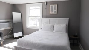 Ein Schlafzimmer mit Bett, Nachttisch, Stuhl, Fenster und einem an der Wand befestigten Spiegel.