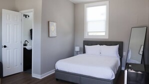 Quarto com cama, janela, porta, espelho e banheiro pequeno com pia.