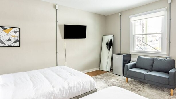 Ein modernes Hotelzimmer mit großem Fenster, Flachbildfernseher, grauem Sofa und einem Bett mit weißer Bettwäsche.