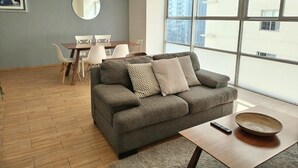 En moderne stue med en grå sofa, et spisebord med stole og et stort vindue med udsigt til bygninger.