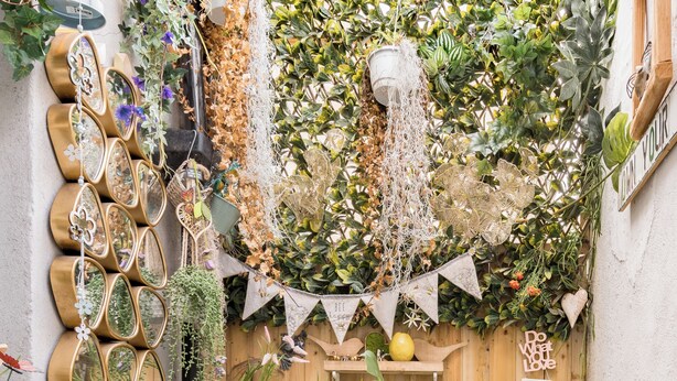 A small patio area with a round table, two blue chairs, a potted plant, and a wooden fence adorned with greenery and decorative items.