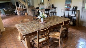 Uma área de refeições com uma grande mesa de madeira, piso com ladrilhos hexagonais e várias cadeiras. Há uma lareira de tijolos ao fundo e uma escadaria que leva a um andar superior.