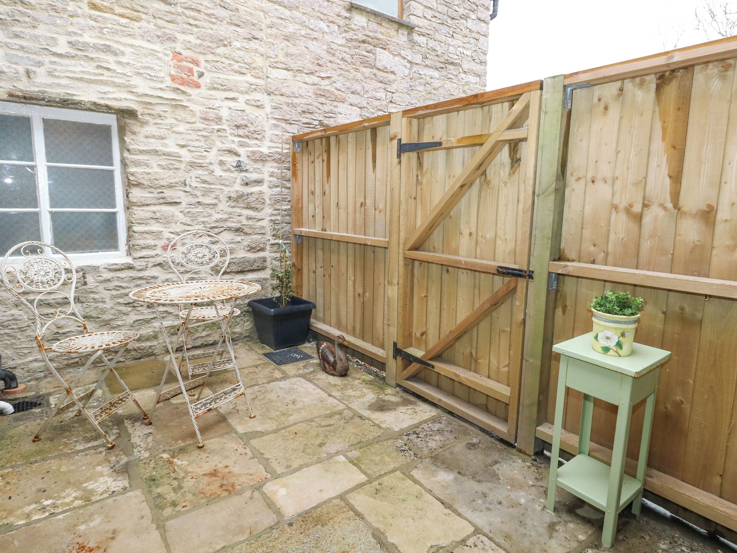 Un bâtiment en pierre avec une porte en bois, une table en métal avec des chaises et une plante en pot posée sur un tabouret vert.