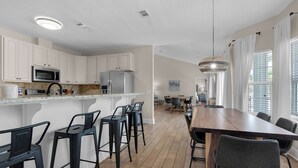 A modern kitchen with white cabinets, a stainless steel refrigerator, and a microwave. There is a dining area with a wooden table and black chairs. - Tranquility 🌴 Huge Pool, Game Room, Sleeps 32, Pet-Friendly, Steps to Beach! (Pensacola Beach)