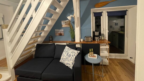 A modern living room with a black sofa, a wooden coffee table, a white staircase, and a kitchen area visible through an open door.