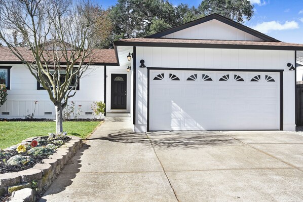 A single-story house with a white garage door, a small front porch, and a tree in the front yard.
