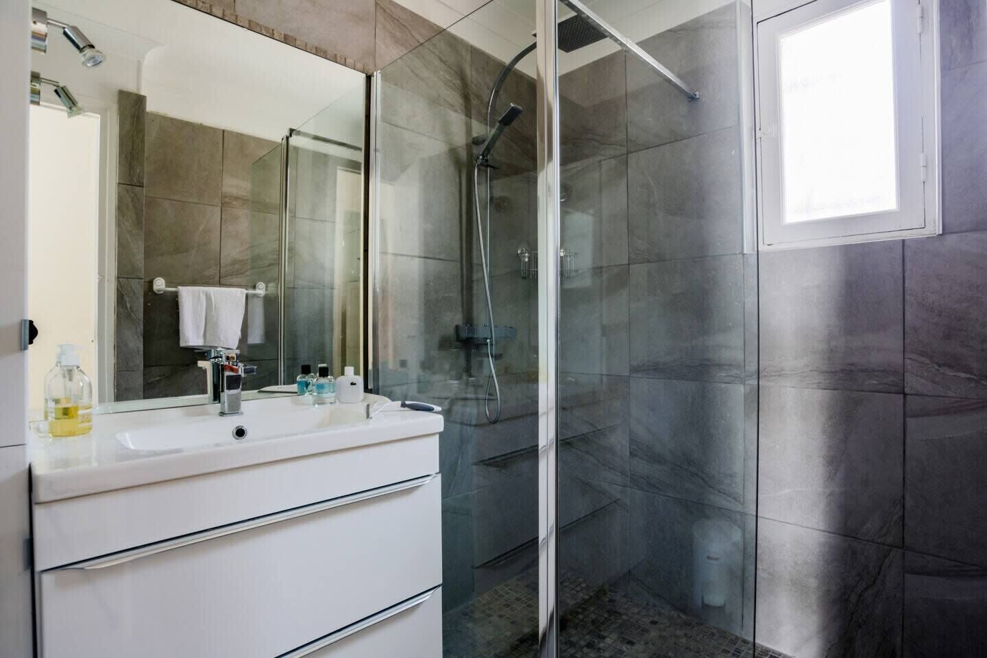 Une salle de bain moderne dotée d’une douche à parois de verre, d’un meuble-lavabo blanc avec un miroir et d’une fenêtre laissant entrer la lumière naturelle.