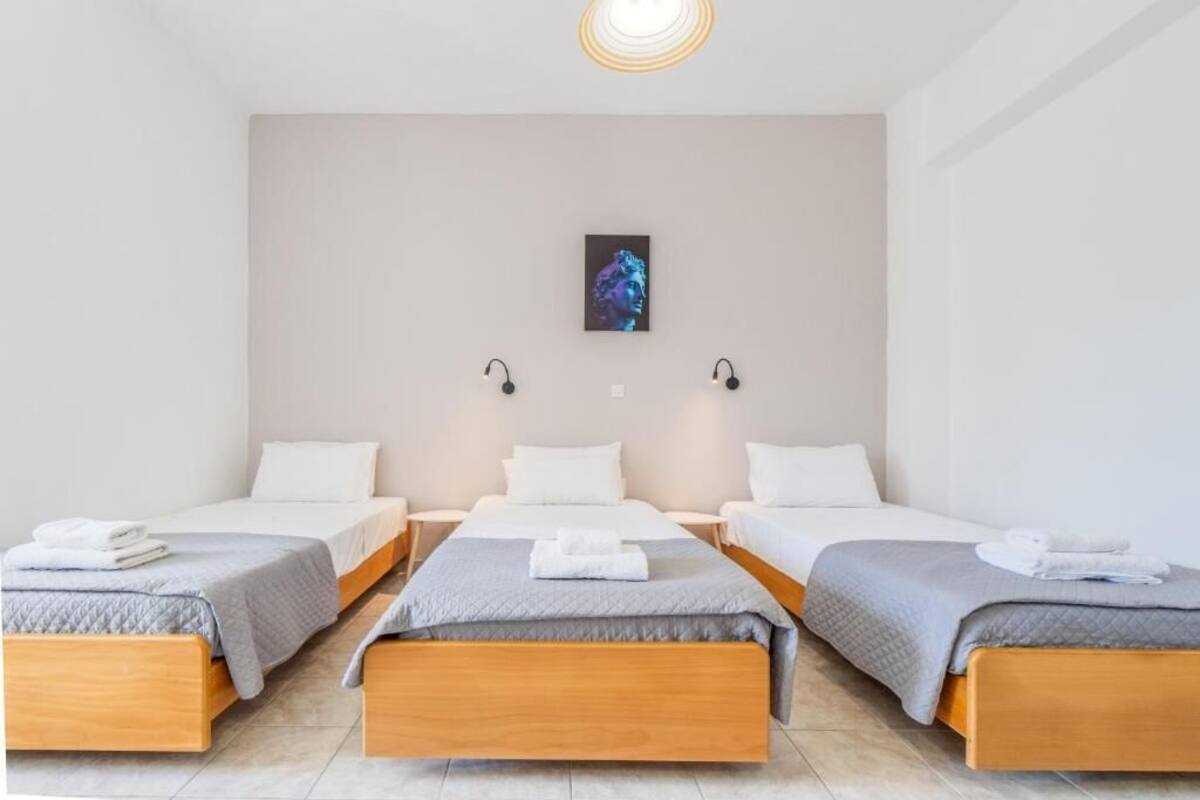 Habitación de hotel con tres camas individuales, cada una con estructura de madera y ropa de cama blanca. Sobre las camas hay una obra de arte colgada en la pared, y dos lámparas de pared a cada lado.