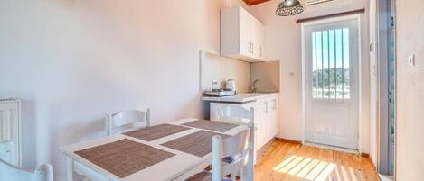 A small, bright kitchen with white cabinets and a wooden ceiling. There is a dining table with chairs and a window providing natural light.