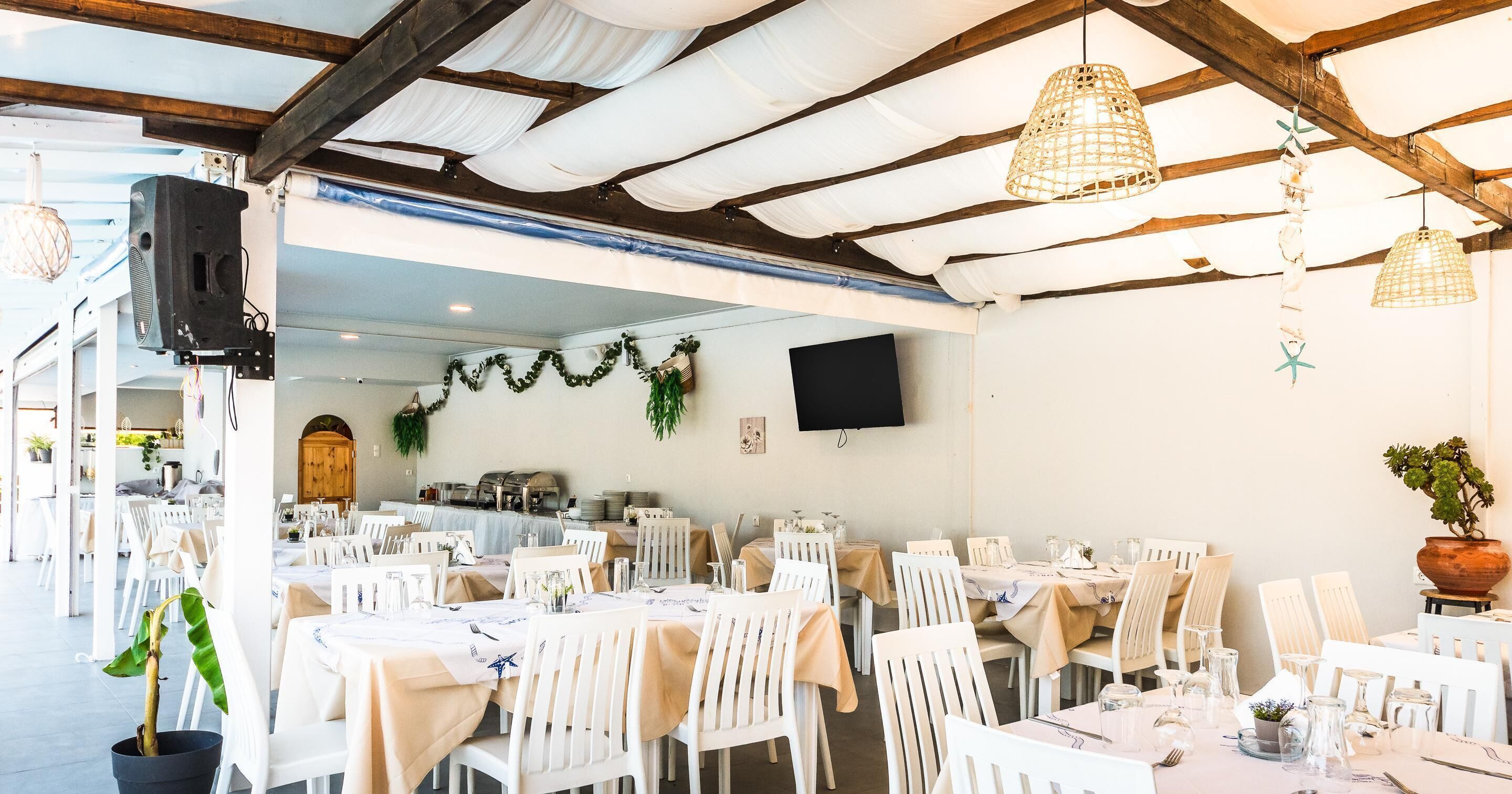 A spacious indoor dining area with white tables and chairs, wooden beams on the ceiling, and hanging pendant lights.