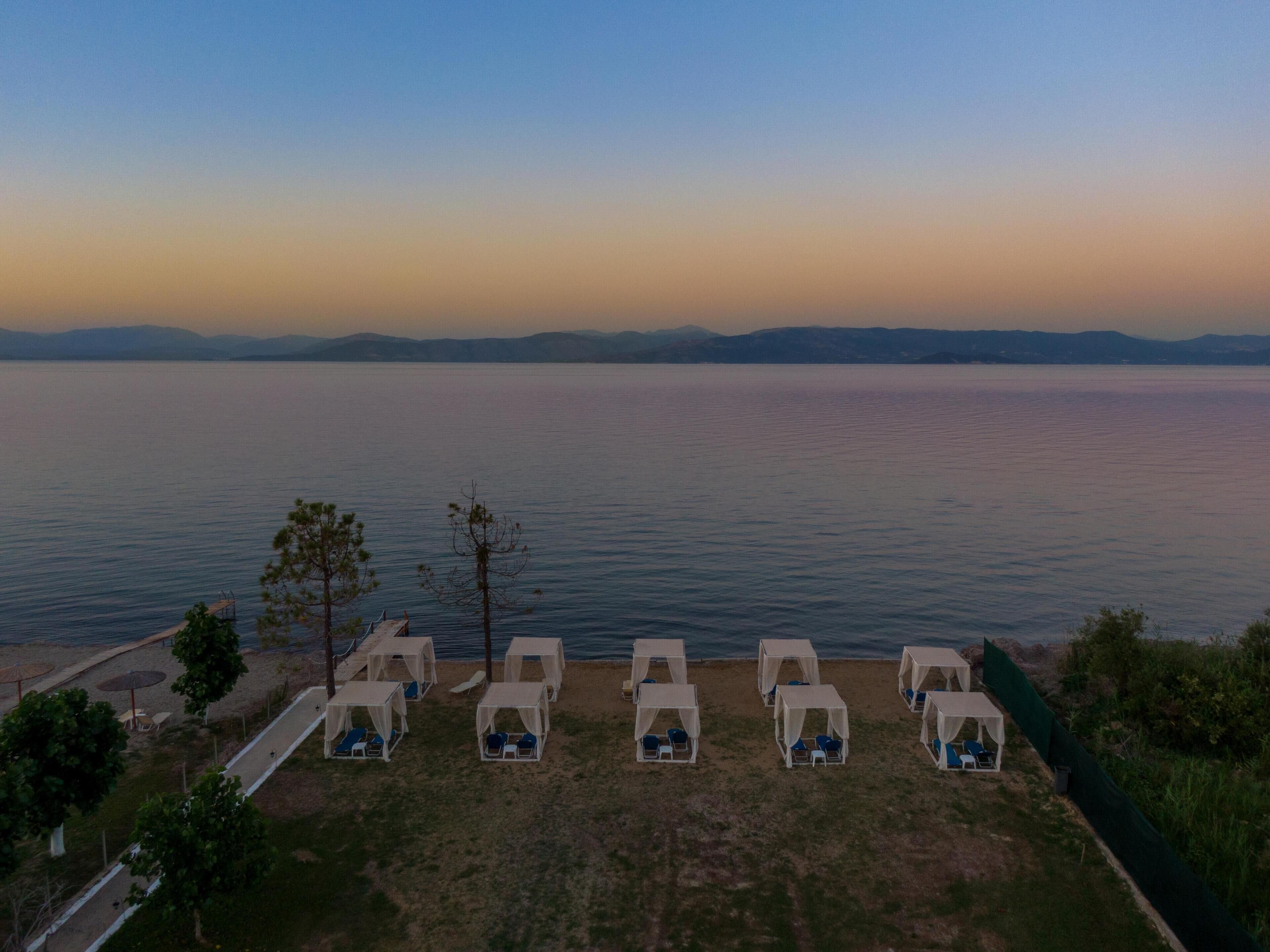 A beachside view with multiple tents and lounge chairs arranged along the shore, overlooking a calm sea and distant mountains.