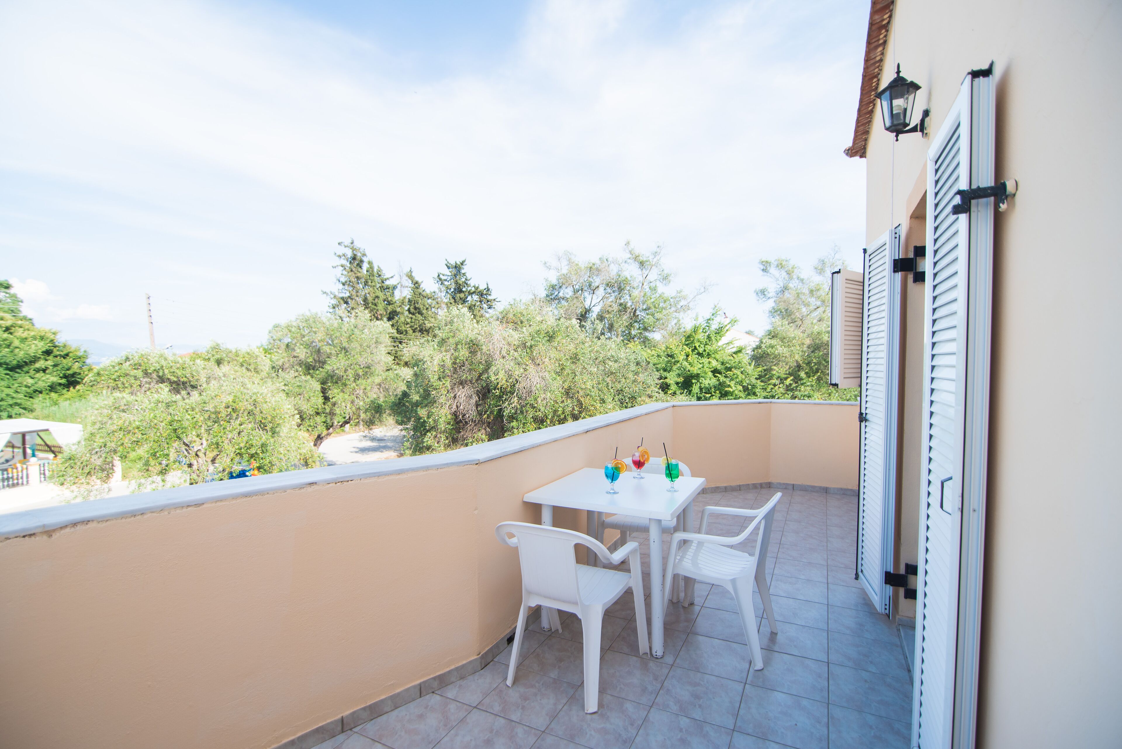 En balkon med et hvidt bord og stole, udsigt til grøn natur, og en bygning med skodder.