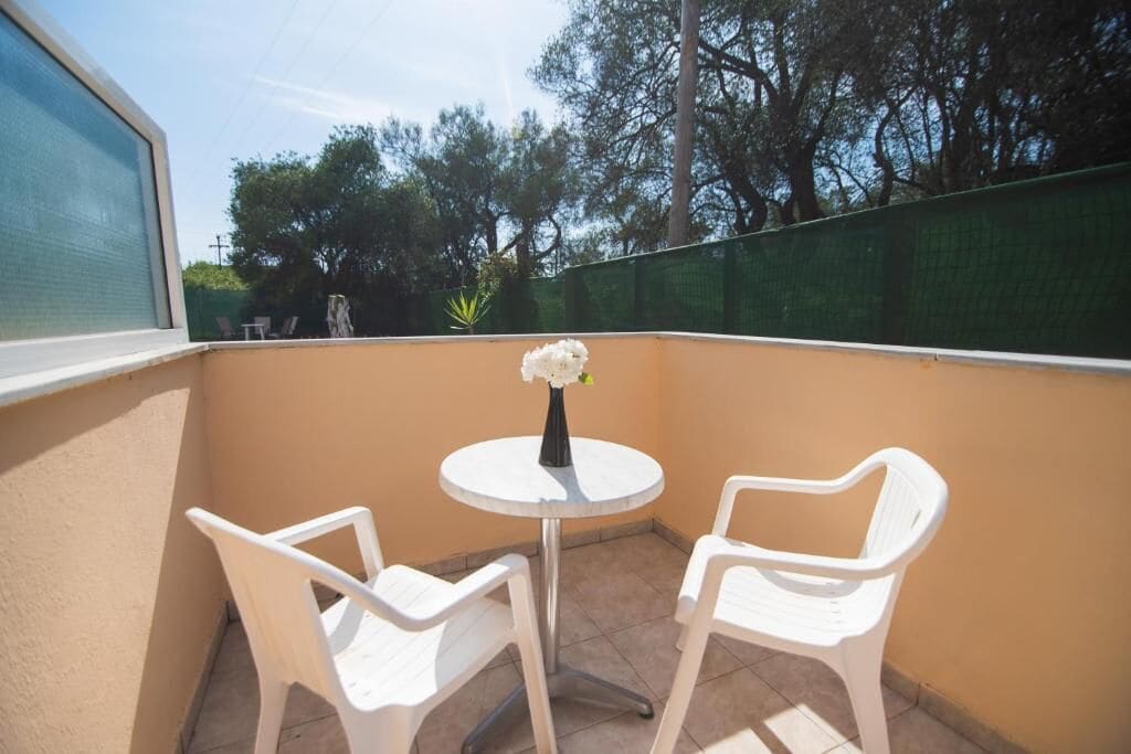 Een klein balkon met twee witte plastic stoelen en een ronde tafel, een vaas met bloemen en uitzicht op bomen en een groene hek.