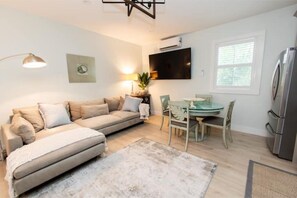 A modern living room with a beige sofa, a dining table with chairs, a flat-screen TV mounted on the wall, and a refrigerator.