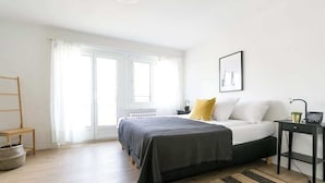 Une chambre à coucher avec un lit, une table de chevet, une chaise, une fenêtre avec des rideaux et un radiateur.