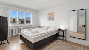 A modern bedroom with a large bed, a bedside table with a lamp, a mirror, and a view of the outdoors through large windows. - Cozy Central Lucerne Apartment with Balcony (Luzern)