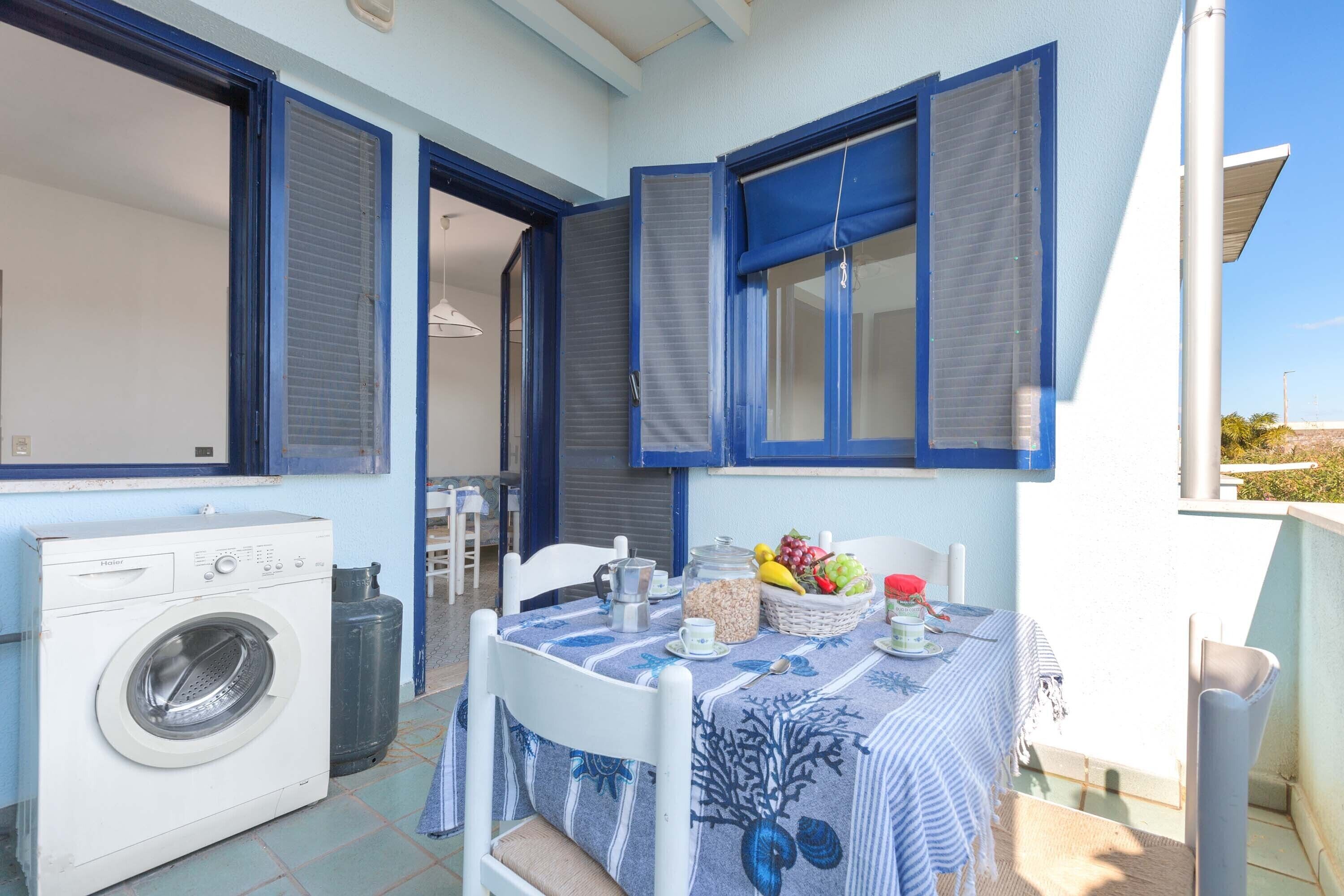 A balcony with a table set for breakfast, a washing machine, and a window with shutters.