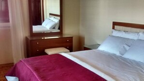 Een opgemaakt bed met een wit en paars dekbed, een houten nachtkastje, een spiegel en een klein bankje.