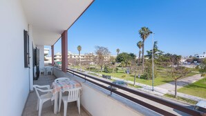 Un balcón con mesa y sillas, con vista a una calle con palmeras y edificios.