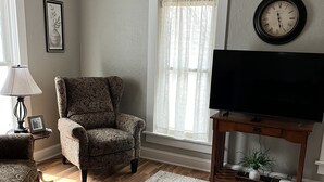 A living room with a patterned armchair, a television on a wooden stand, a white rug, and a window with sheer curtains. - The Krause Historic Eau Claire Home (Eau Claire)