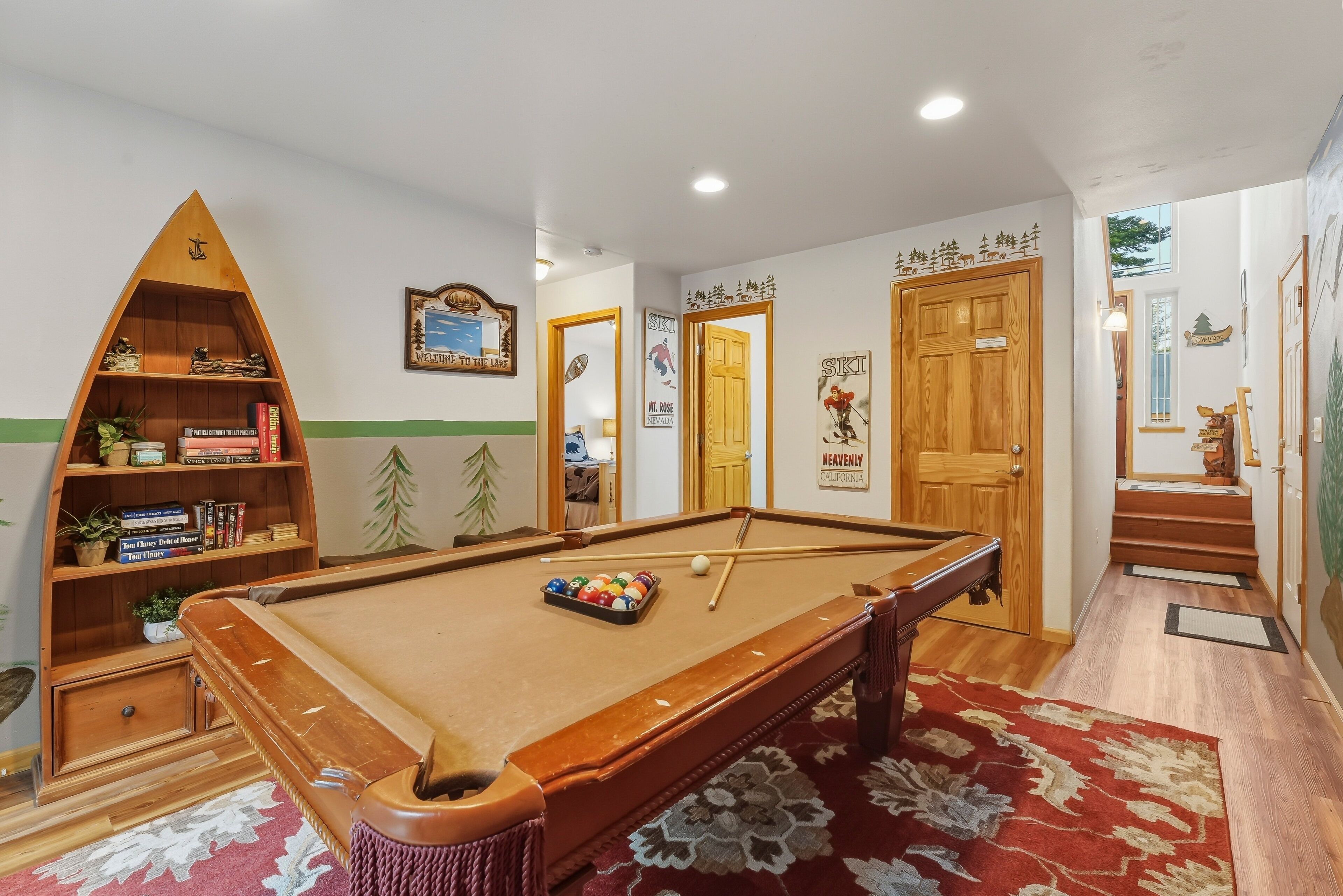 A pool table with a red patterned rug, a wooden bookshelf with books and plants, and a door with a ski-themed sign.