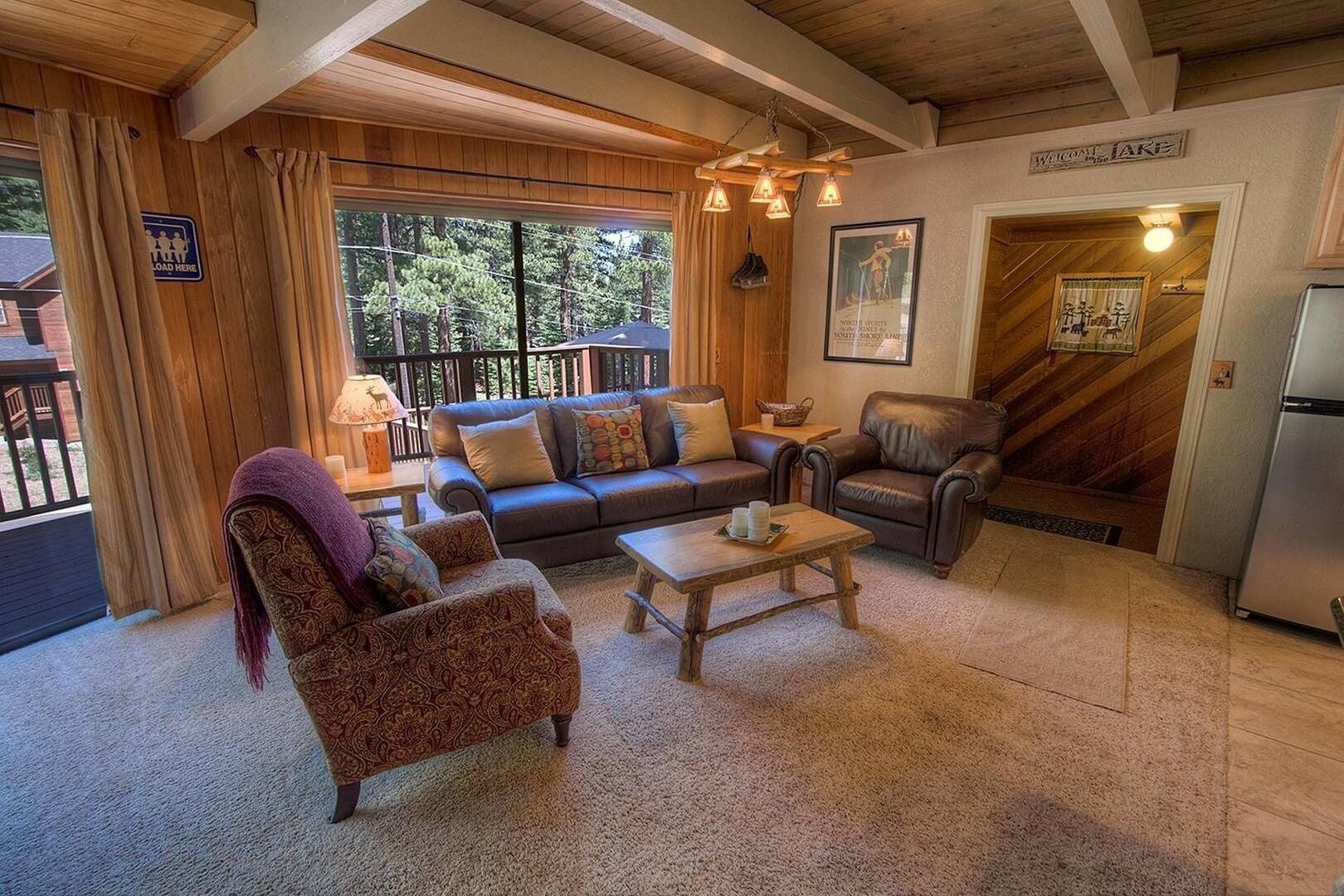 A cozy living room with a sofa, armchair, and coffee table, wooden ceiling beams, and a view of the outdoors.