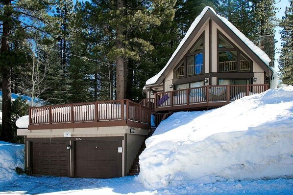 Una cabaña con un garaje amplio y una terraza cubierta, rodeada de nieve y árboles.