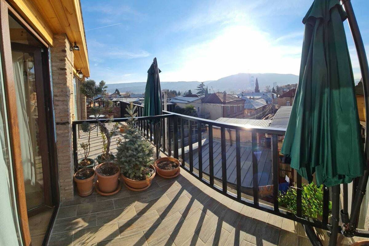 Un balcon agrémenté de plantes en pot, un parasol vert et une vue sur un paysage urbain avec des montagnes en arrière-plan.