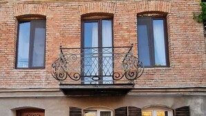 A two-story building with a balcony featuring wrought iron railings. There are three windows on the upper floor and two on the lower floor, each with shutters. - ApartTen (Tbilisi)