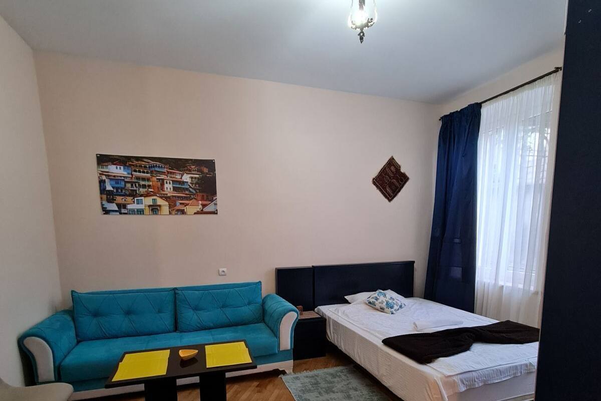 Une petite pièce bien éclairée, avec un canapé bleu, un lit, une petite table sur laquelle se trouvent des dossiers jaunes, un luminaire fix é au mur et une affiche représentant une ville côtière.