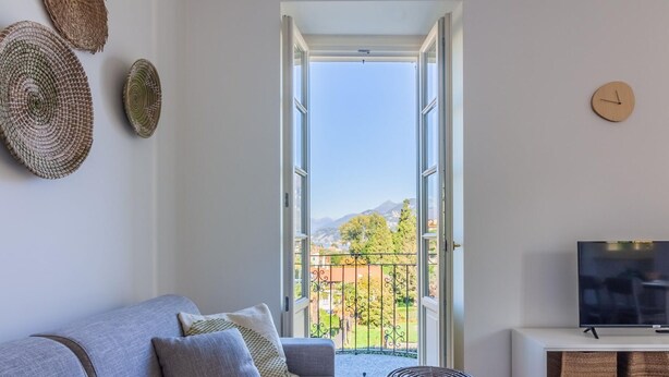 A living room with a sofa, a TV, and a view of a balcony with a mountain backdrop. - Green Rose by Wonderful Italy (Bellagio)