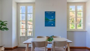 A dining area with a table and chairs, a poster of Como Lake, and large windows with a view of buildings. - Green Rose by Wonderful Italy (Bellagio)