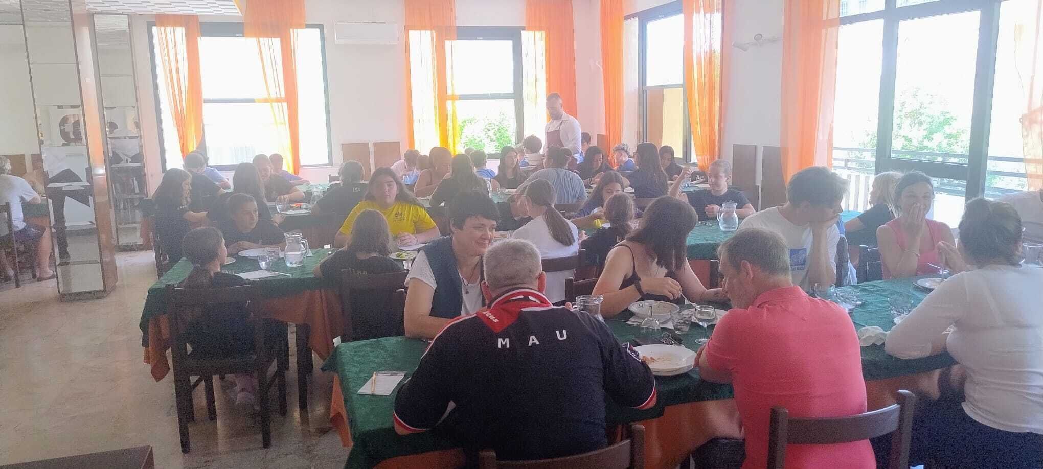 A dining area with multiple tables and chairs, large windows with orange curtains, and a person standing at the front.