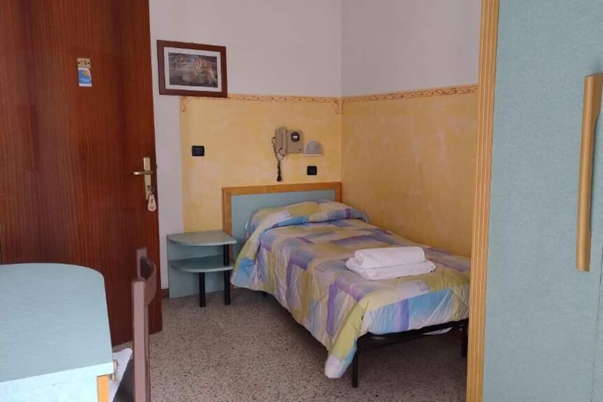 A small, single-bed room with a wooden headboard, a bedside table, and a chair. There is a wardrobe with two doors and a small wall-mounted telephone.
