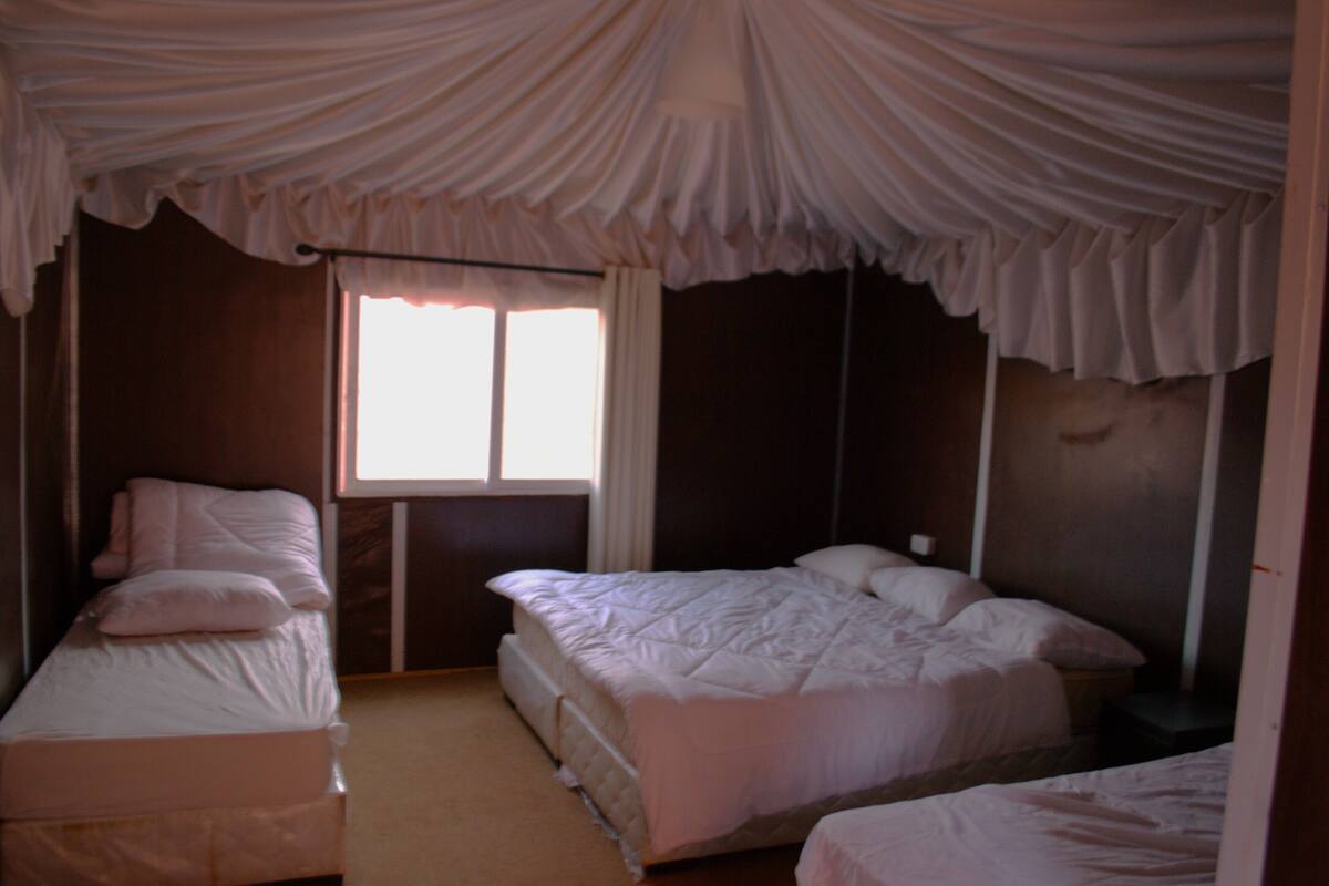 Quarto com duas camas de solteiro, uma janela grande com cortinas e teto com tecido drapeado.
