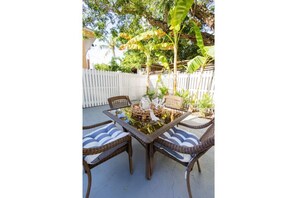 Une terrasse extérieure dotée d’une table à plateau de verre et de chaises en osier, entourée d’une clôture blanche et de plantes tropicales.