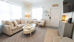 A living room with a beige sofa, a round coffee table, and two armchairs. There is a television mounted on the wall and a cabinet with a decorative item on top. - The Sandy Beach House in Historic Key West (Key West)