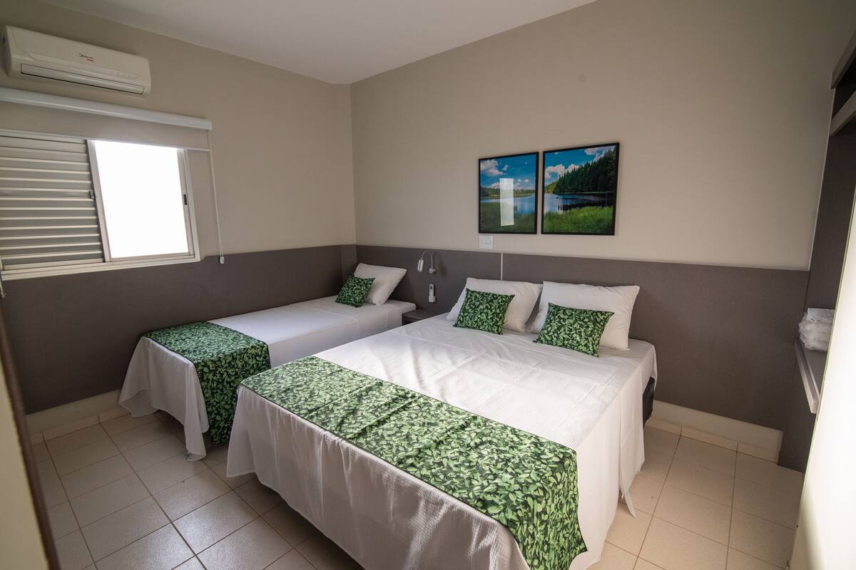 A hotel room with two beds, a window with blinds, an air conditioner, and two framed pictures on the wall.