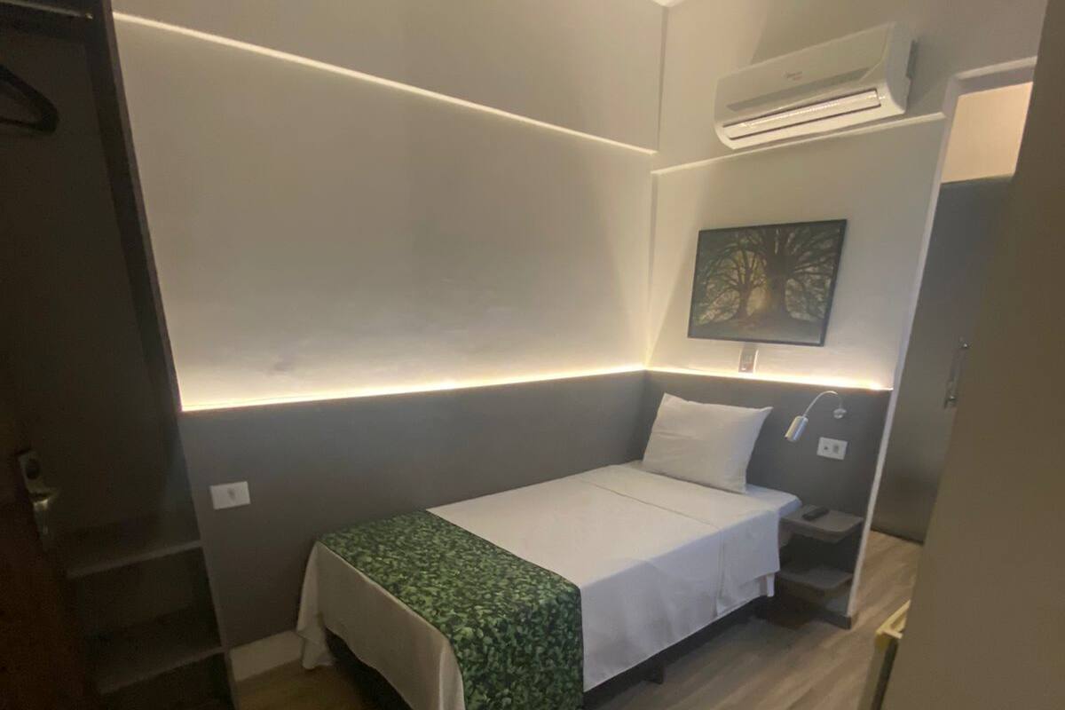 A small, compact room with a single bed, a bedside table, and a wall-mounted air conditioning unit.