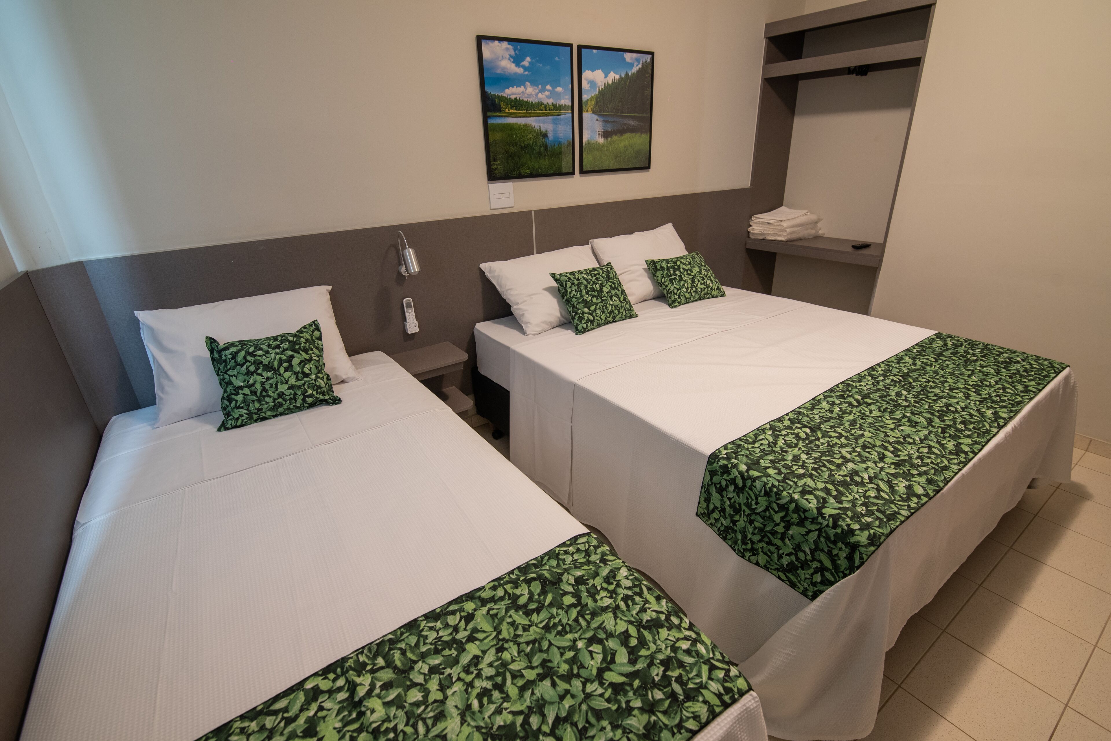 A hotel room with two beds, each with white bedding and green leaf-patterned throw pillows. There are two framed pictures on the wall, a nightstand with a lamp, and a shelf with folded towels.