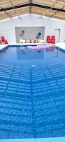 An indoor swimming pool with clear blue water, surrounded by red and white lounge chairs.