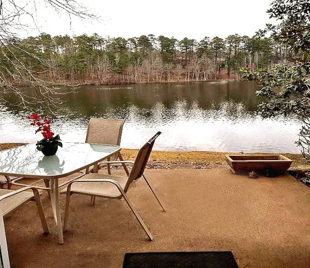 Een terras met een glazen tafel en stoelen, uitkijkend op een meer, omringd door bomen.